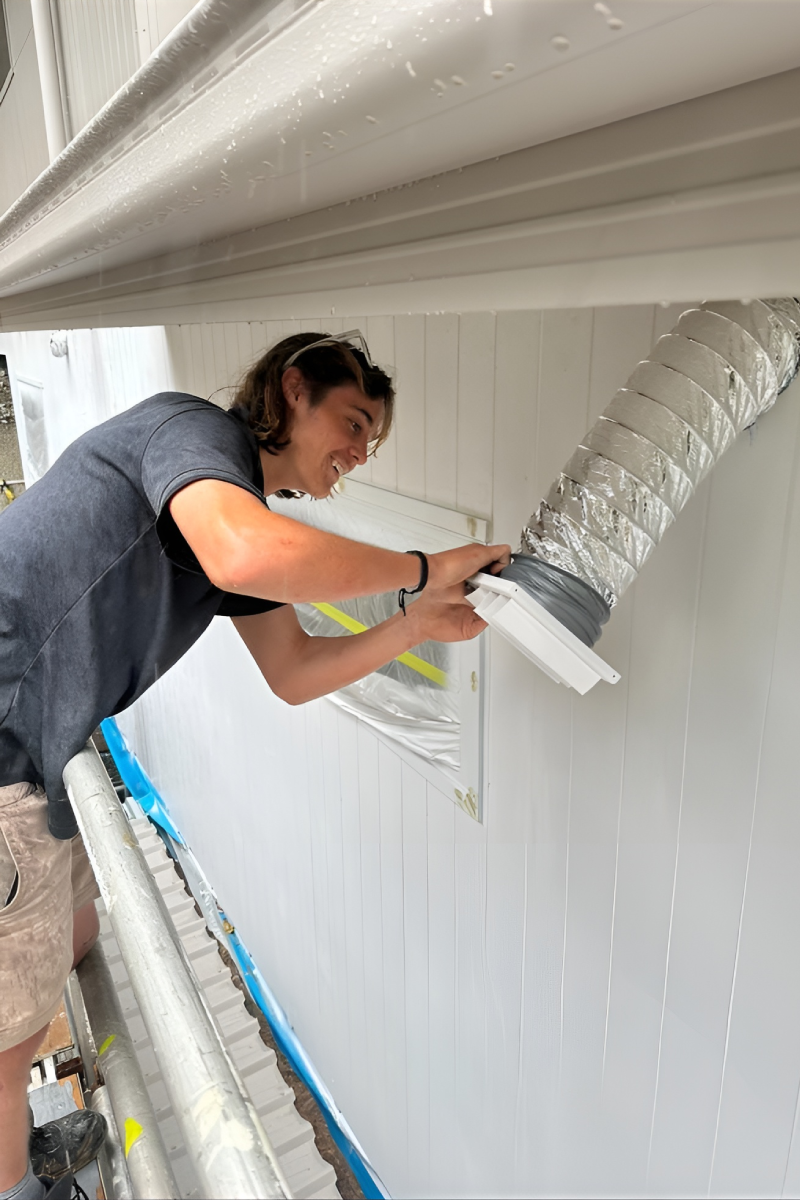 Person installing an air duct vent on a white exterior wall, standing on scaffolding for support.