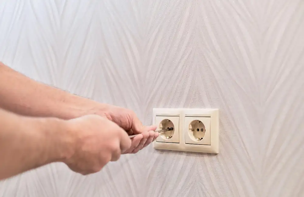 Hands use a screwdriver to adjust or repair a wall-mounted double electrical outlet on a patterned wallpaper background.