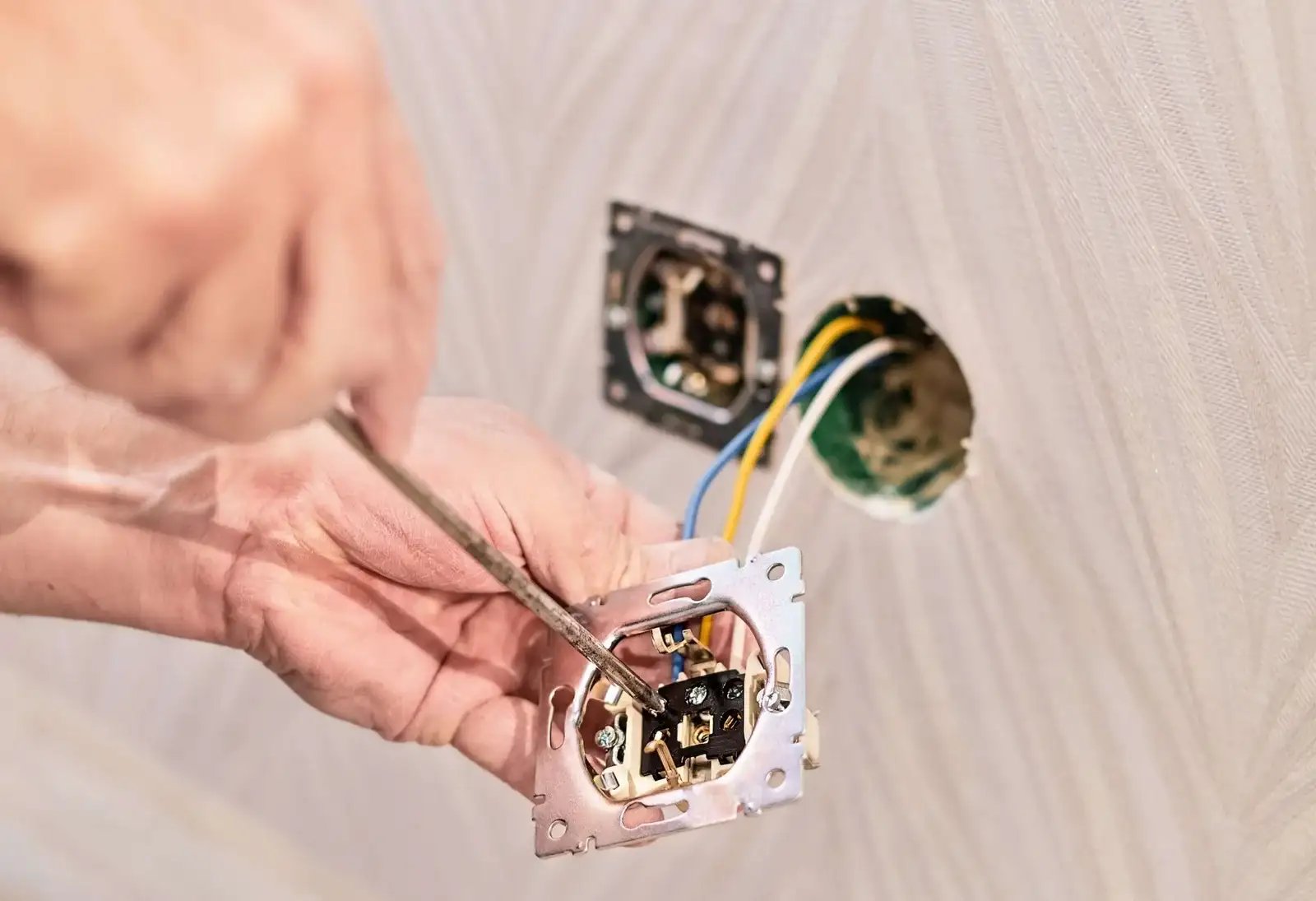 Hands using a screwdriver to wire an electrical outlet in a wall, with exposed wires and another outlet box visible.