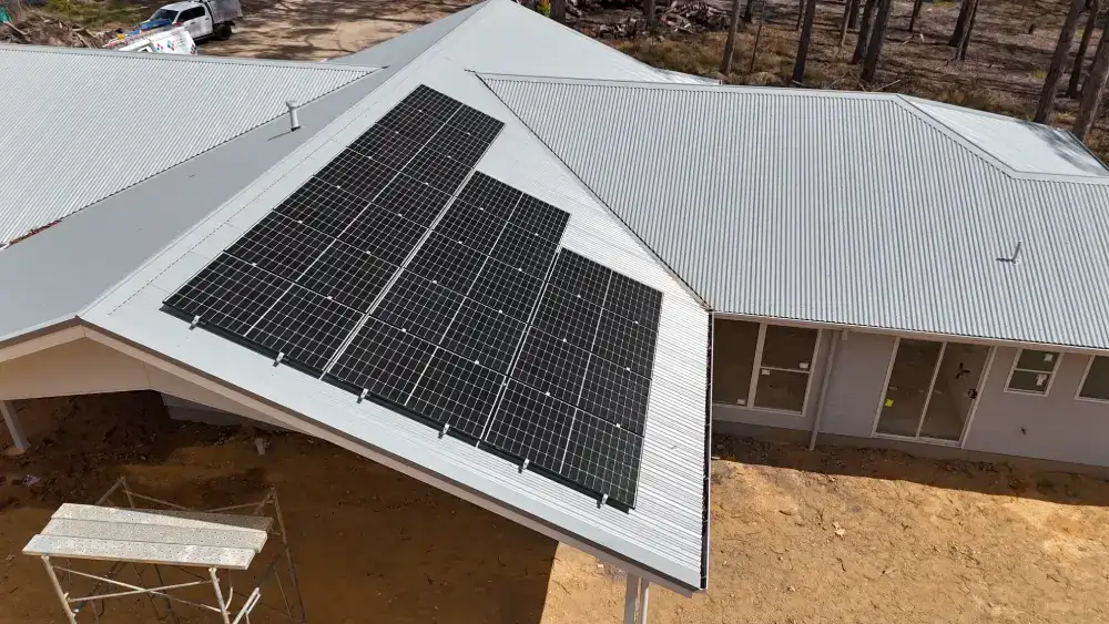 Aerial view of a house with solar panels installed on a metal roof, surrounded by a forested area and sandy ground.