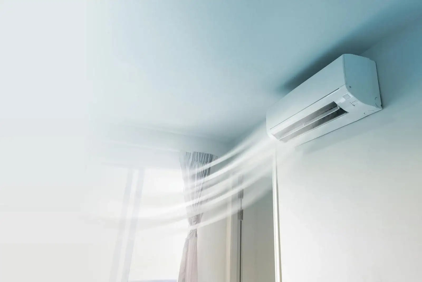 Wall-mounted air conditioner blowing cool air in a bright room with a window and light curtain.