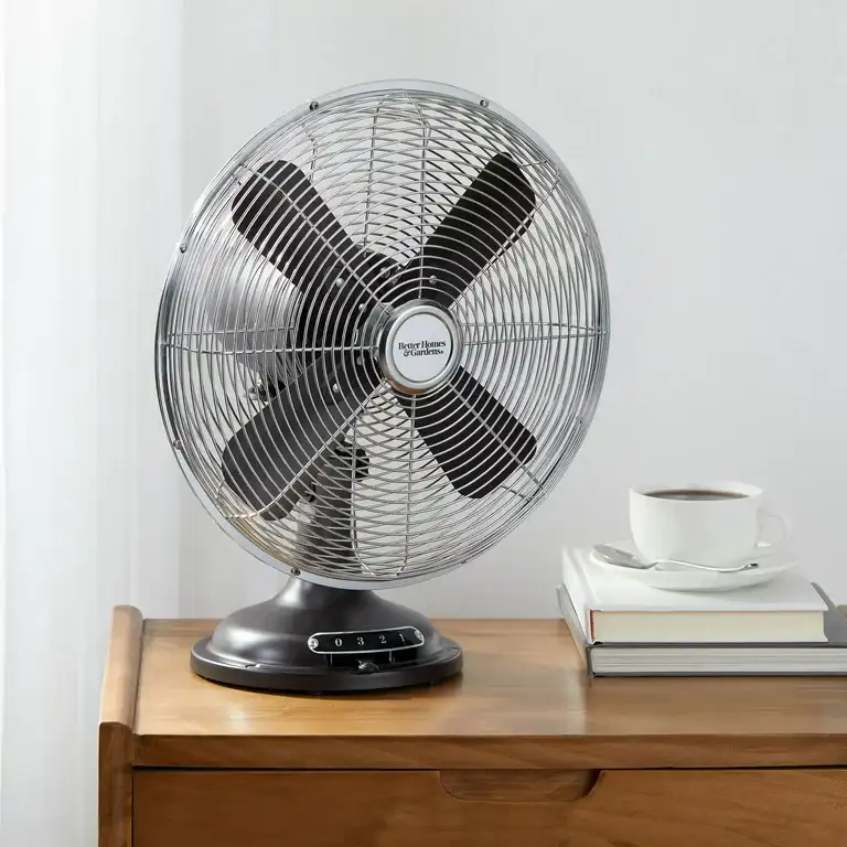 A metal desktop fan on a wooden table beside stacked books and a white coffee cup.