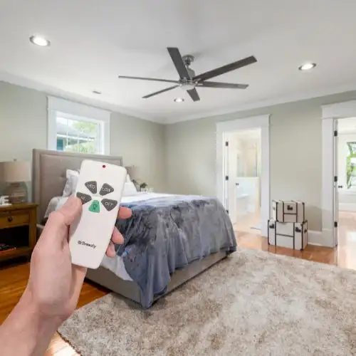 Hand holding a ceiling fan remote in a modern bedroom with a large bed, wooden floor, and white doors.