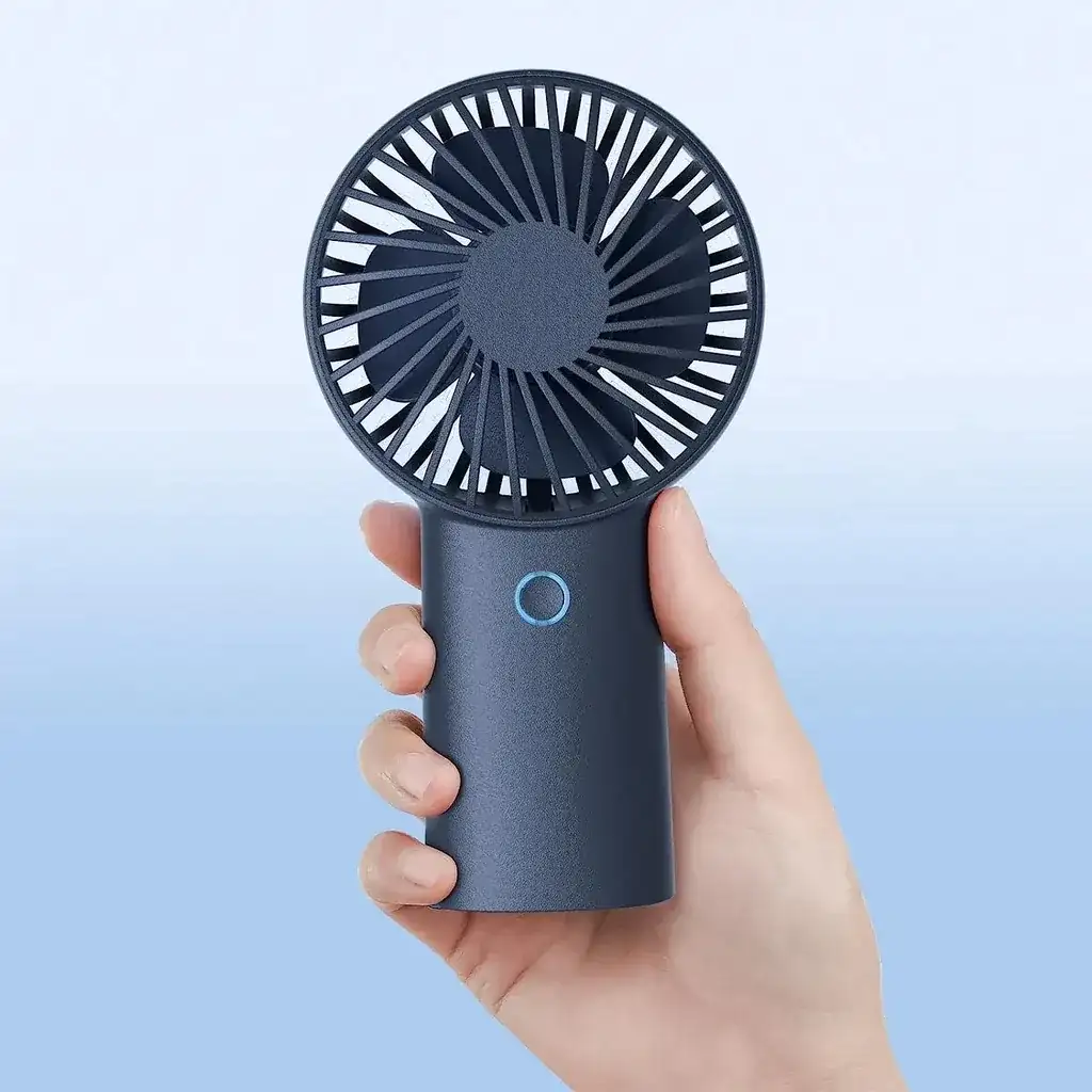 A hand holding a small, black portable fan with a circular blue button on the handle, against a gradient blue background.