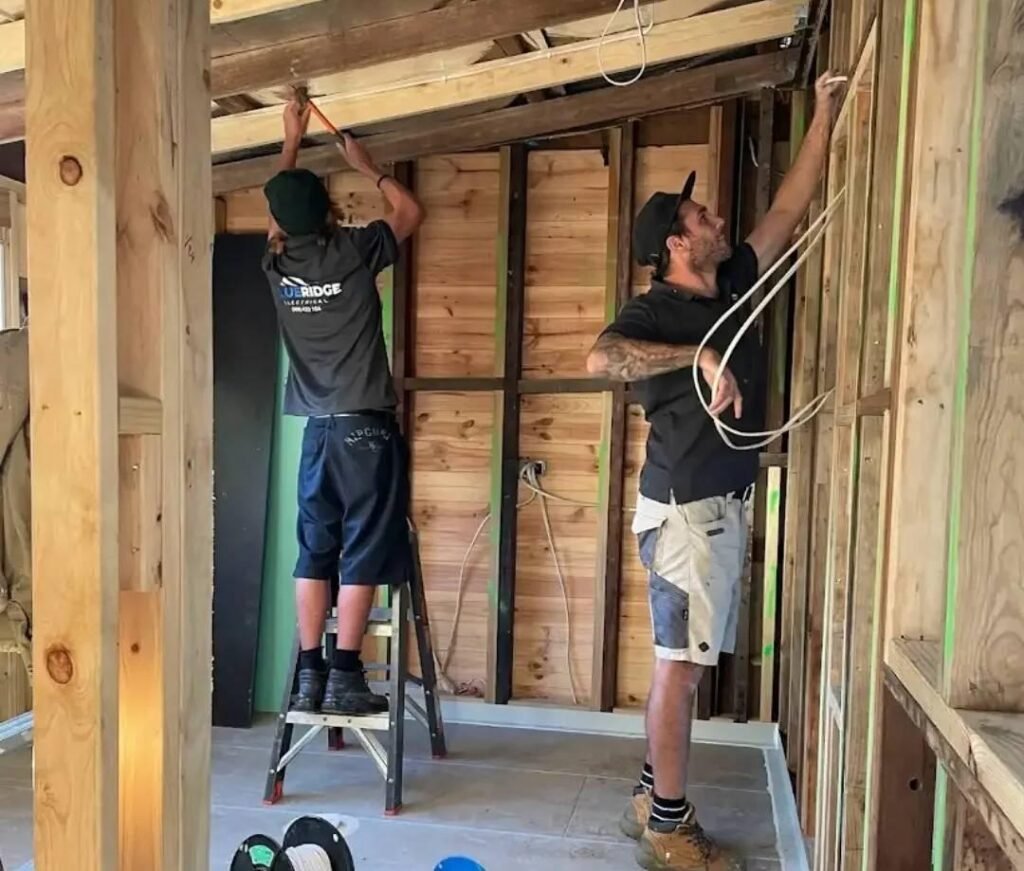 Two electricians installing wiring in a timber-framed house under construction, with one on a step ladder working on the ceiling and the other organizing cables along the wall frame.