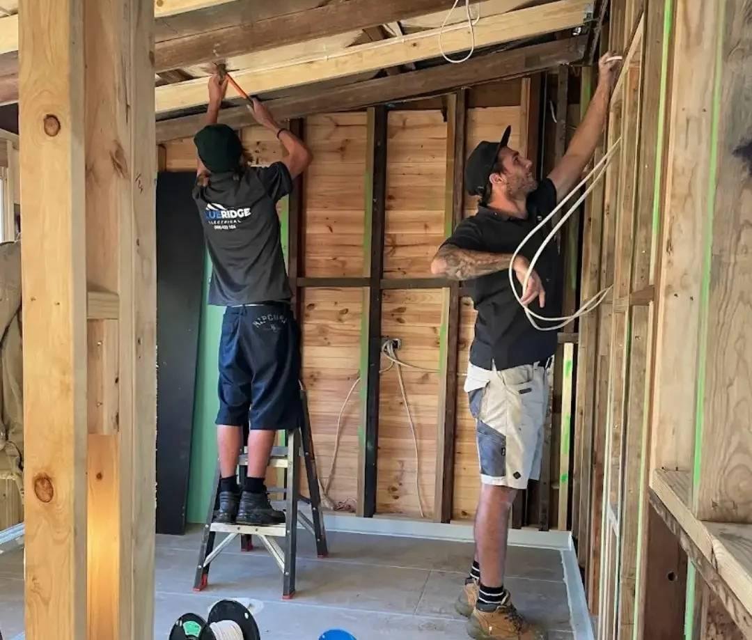 Two electricians installing wiring in a timber-framed house under construction, with one on a step ladder working on the ceiling and the other organizing cables along the wall frame.