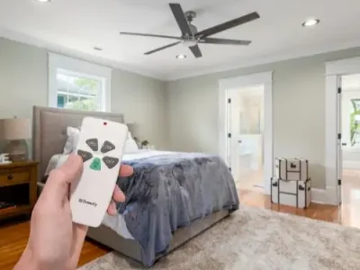 Hand holding a ceiling fan remote in a modern bedroom with a large bed, wooden floor, and white doors.