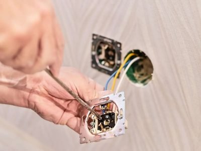 Hands using a screwdriver to wire an electrical outlet in a wall, with exposed wires and another outlet box visible.