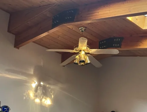 A ceiling fan with gold fixtures and wooden blades is mounted on a wooden ceiling, casting shadows on the wall.